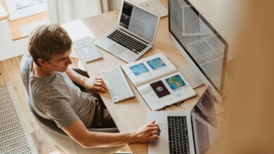 A man multitasks with laptops and a desktop, coding in a home office setting.